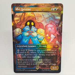 Può includere: Una carta collezionabile con Vivi Ornitier, una Creatura Leggendaria Mago. La carta presenta una vivace illustrazione di un personaggio in abito giallo e blu. Il testo della carta include le istruzioni di gioco e un potere di 0/3.