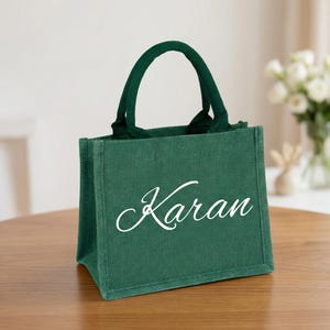 May include: A green jute tote bag with a handle. The bag has the name "Karan" in white script across the front. The bag is sitting on a wooden table.