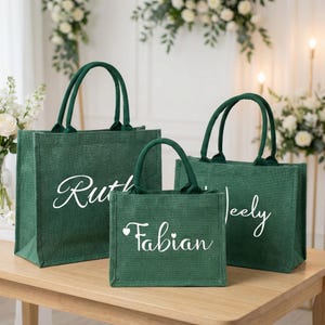 May include: Three green jute tote bags with white script names: "Ruth", "Fabian", and "Jeely". The bags have matching green handles and are arranged on a light brown wooden table. The background includes floral arrangements.