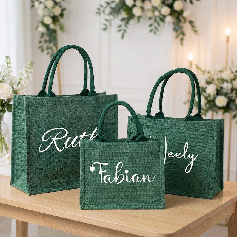 May include: Three green jute tote bags with white script names: "Ruth", "Fabian", and "Jeely". The bags have matching green handles and are arranged on a light brown wooden table. The background includes floral arrangements.