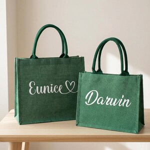 May include: Two green burlap tote bags with matching green handles. The larger bag has the name "Eunice" in white script with a heart symbol. The smaller bag has the name "Darwin" in white script.