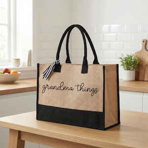 Peut inclure: Un sac fourre-tout rectangulaire avec des poignées et des bordures noires. Le sac est fait d'un matériau en toile de jute beige avec une section inférieure noire. Les mots "grandma things" sont écrits en cursive sur le devant.