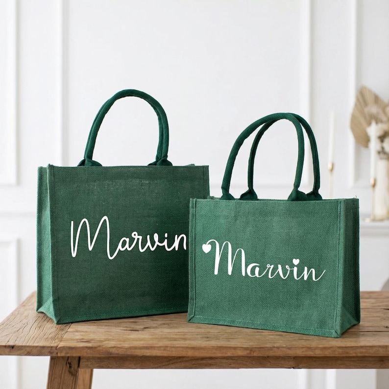 May include: Two green jute tote bags with green handles. The larger bag has the name "Marvin" in white script. The smaller bag also has "Marvin" in white script, with a small heart symbol. Both bags are on a wooden surface.