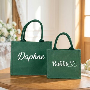 May include: Two green tote bags with green handles. The larger bag has the name "Daphne" in white script, and the smaller bag has the name "Babbie" with a heart symbol. The bags are on a wooden surface.