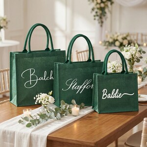May include: Three green tote bags with white cursive text, displayed on a wooden table with a white runner. The bags have matching green handles. The bags are different sizes and have the names "Balch", "Staffor", and "Baldie" printed on them.