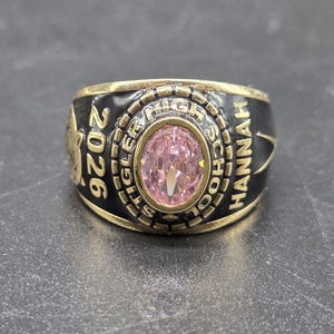May include: A gold-toned class ring with a black band and a large, oval pink gemstone. The ring is inscribed with "2026", "Stigler High School", and "Hannah". The ring has a classic, bold design.