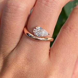 May include: A rose gold ring featuring a pear-shaped diamond. The ring's band curves gracefully, cradling the sparkling gemstone. The diamond is clear and faceted, catching the light. The ring is shown on a hand.