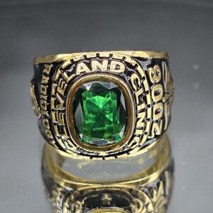 May include: A gold-toned ring with a large, emerald-green gemstone at its center. The ring's band features black enamel with the words "RADIOLOGY AND CLINIC 2008" in raised gold lettering. The ring is highly reflective.