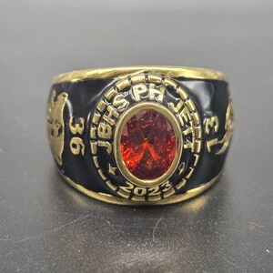 May include: A gold and black class ring with a large, oval, red gemstone at the center. The ring has the text "JBHS PH JEFF 2023" and the numbers "36" and "13" on the sides. The ring is likely a high school or college class ring.