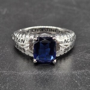 May include: Silver ring with a large, rectangular, dark blue gemstone. The ring has engraved text around the band, including "IDE CHRISTINE HU" and "ACADE 2026". The ring is set against a dark background.
