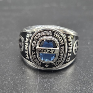 May include: Silver-toned class ring with a blue gemstone center. The ring is inscribed with "WEST FELICIANA HIGH SCHOOL," "SAINTS," "ETHAN," and the year "2027."