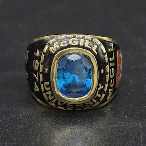 May include: A gold-toned ring with a large, oval, blue gemstone at the center. The ring has a black band with the words "McGill University" and the year "1974" inscribed. The ring is a class ring.