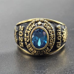 Könnte beinhalten: Ein goldfarbener Ring mit einem großen, ovalen, blauen Edelstein in der Mitte. Der Ring hat schwarze Details mit den Worten "2025", "GEORGE ROGERS CLARK" und "GLORIA" um das Band herum.