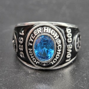 May include: A silver-toned class ring with a large, oval, blue gemstone at the center. The ring features the text "JOHN TYLER HIGH SCHOOL" and "1986". The words "LOVE GOD" are also visible.