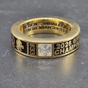 May include: A gold-colored ring with a square, clear gemstone at the center. The ring has black enamel detailing with the text "2026 GW CHAMPION" and "#33". The inside of the ring has the text "JEDEN S INDALECIO JEREMIAH 29:11".