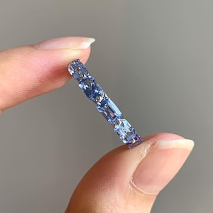 May include: A delicate ring featuring a series of light blue rectangular and square-cut gemstones. The ring is held between two fingers, showcasing the intricate design and the sparkle of the stones. The ring appears to be made of a silver or platinum metal.
