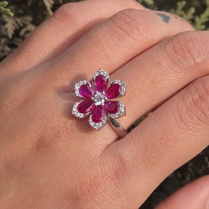 May include: A silver ring featuring a flower design. The flower has six petals of deep red gemstones, each outlined with small, clear crystals. A single, clear gemstone sits in the center of the flower.