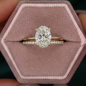 Könnte beinhalten: Ein oval geschliffener Diamant-Verlobungsring und ein Diamantband liegen in einer rosa sechseckigen Ringschatulle. Die Ringe sind goldfarben. Der Hintergrund ist ein dunkelgrüner Stoff.