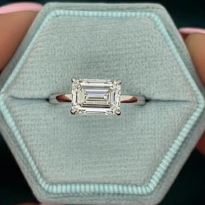 Può includere: Un anello di fidanzamento in argento con diamante taglio smeraldo è esposto in un portagioie di velluto azzurro chiaro. L'anello è tenuto da una persona con lo smalto rosa. Lo sfondo è di colore verde acqua scuro.