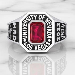 Peut inclure: Bague en argent avec une pierre rouge rubis rectangulaire au centre. La bague porte les inscriptions "UNIVERSITY OF NEVADA LAS VEGAS 2022" et "HRD". Elle est posée sur une surface en marbre blanc.
