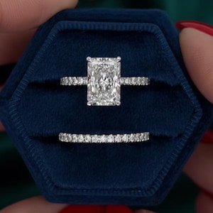 Può includere: Un set di anelli di fidanzamento color argento con una grande pietra centrale rettangolare taglio radiante e una fede pavé abbinata, entrambi incastonati con piccole pietre chiare, presentati in un cofanetto esagonale in velluto blu scuro.