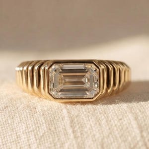 May include: A gold ring featuring a central emerald-cut clear gemstone set within a band characterized by a series of vertical, parallel ridges that extend around the circumference of the ring, creating a textured, ribbed appearance.