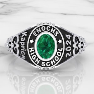 May include: A silver high school class ring with a black band and a green oval-shaped stone. The ring is inscribed with "ENOCHS HIGH SCHOOL 2024" and "Kaprice". The ring is displayed on a white marble surface.