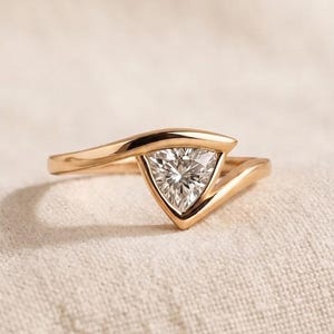 May include: A gold ring featuring a single trillion-cut clear gemstone set within a bypass-style band. The metal has a polished finish, and the triangular stone is held securely by the sweeping, asymmetrical arms of the ring's shank.