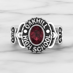 May include: A silver-colored class ring with a large oval, dark red stone at the center. The ring features the words "OAKHILL HIGH SCHOOL" in black lettering around the stone. The year "2021" and the initials "AJE" are also visible on the ring.