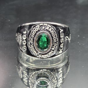 May include: A silver-toned class ring with a large, oval, emerald-green gemstone at its center. The ring is engraved with the words "High School", "Kasey", and the year "1999". The ring is highly reflective.