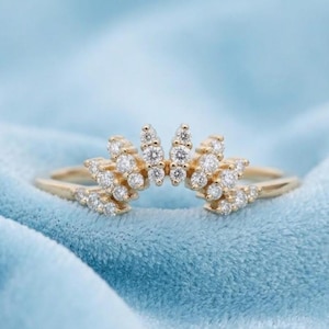 May include: A gold-colored ring with a crown-like design. The ring features multiple small, clear gemstones set in a curved pattern. The ring is set against a light blue background.