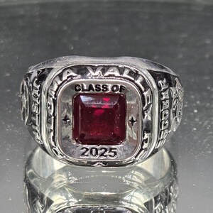 May include: A silver-toned class ring with a square, deep red stone at the center. The ring is engraved with the words "CLASS OF 2025" and other text around the band. The ring is highly reflective.