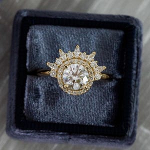 May include: A gold engagement ring with a large, round, clear central stone, encircled by smaller stones in a halo design. The ring is presented in a dark blue velvet box. The ring's design features a sunburst-style halo, adding to its elegance.