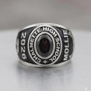 May include: A silver-toned class ring with a dark oval-shaped stone at the center. The ring is inscribed with "CHALMETTE HIGH SCHOOL" and "2026 MOLLIE". The ring has a black border.