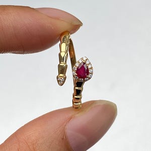 Könnte beinhalten: Ein goldfarbener Ring in Schlangenform mit einem birnenförmigen, tiefroten Edelstein, umgeben von kleinen, klaren Steinen. Der Ring hat ein segmentiertes Design am Körper und einen kleinen, klaren Stein am Kopf.