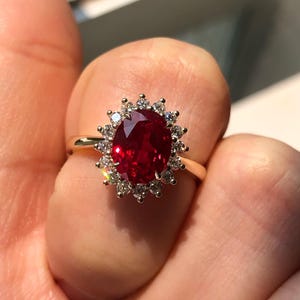 May include: A gold ring with a large, oval-cut, deep red gemstone, encircled by a halo of smaller, clear gemstones. The ring's band is plain gold. The ring is held up by a person's fingers.