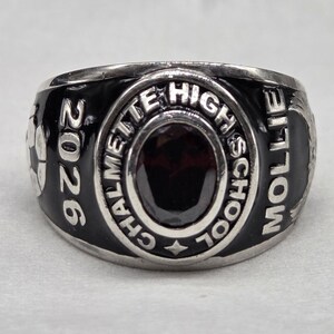 May include: A silver-toned class ring with a black enamel band. The ring features the text "CHALMETTE HIGH SCHOOL" and "2026". The center stone is dark red. The name "MOLLIE" is also inscribed on the ring.