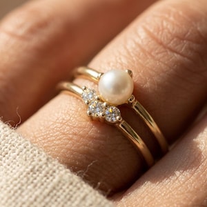 Peut inclure: Deux bagues en or, l'une avec une grosse perle et l'autre avec trois petits diamants. Les bagues sont empilées sur un doigt, mettant en valeur le design délicat et le savoir-faire de la bijouterie.