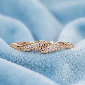 May include: A gold-colored ring with a flowing, intertwined design. The ring is adorned with small, sparkling gemstones set along the curves. The ring is set against a soft, light blue background.