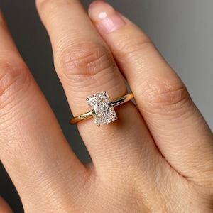 Può includere: Un anello con fascia in oro e gemma rettangolare trasparente. La gemma è incastonata in una fascia d'oro. L'anello è mostrato su un dito.