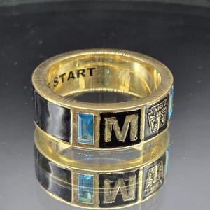 May include: A gold-toned ring with black enamel detailing. The ring features the word "START" engraved on the inside. The exterior has the letters "M" and "W" in black enamel, with blue rectangular accents and small symbols.