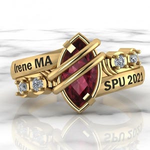 May include: A gold ring with a marquise-cut, deep red gemstone at the center. The ring features the inscription "(irene MA)" on one side and "SPU 2021" on the other. Small, clear gemstones are set on either side of the central stone.