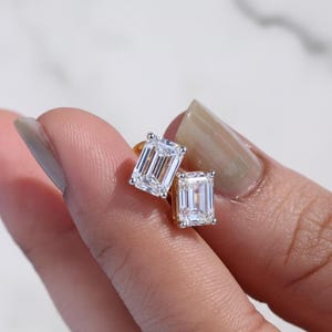 May include: A pair of emerald-cut diamond stud earrings. The diamonds are set in a gold setting. The earrings are displayed against a neutral background.