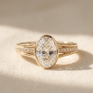 May include: A gold ring with a large, oval-cut diamond as the centerpiece. The band features a row of smaller diamonds, adding sparkle. The ring is set against a neutral background, highlighting its elegant design.
