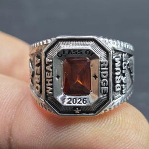 May include: A silver-toned class ring with a rectangular, reddish-brown gemstone at the center. The ring is engraved with the words "CLASS OF 2026", "WHEAT RIDGE", "AERO", and "DRUM MAJOR".