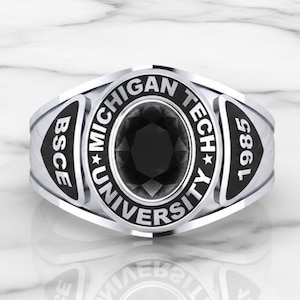 May include: A silver-colored ring with a large, oval black stone at the center. The ring features the words "MICHIGAN TECH UNIVERSITY" and "BSCE" and the year "1985". The ring is set against a marble background.