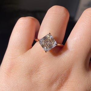 May include: A rose gold ring featuring a large, square-cut, clear gemstone. The gemstone has a multi-faceted design, reflecting light. The ring is set on a thin band, showcasing the prominent stone.