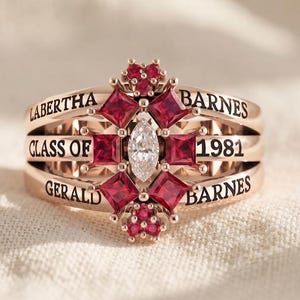 May include: A rose gold ring featuring a central marquise-cut clear stone surrounded by square-cut red stones and small round red accents. The band is engraved with the text LABERTHA BARNES, CLASS OF 1981, and GERALD BARNES in black lettering.