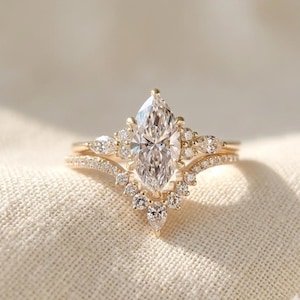 May include: A gold ring set featuring a central marquise-cut diamond flanked by smaller round and pear-shaped diamonds. The set includes a curved band adorned with a row of round diamonds that follows the contour of the main marquise stone.
