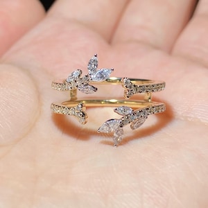 Può includere: Un anello color oro con un design a doppia fascia. L'anello presenta pietre preziose chiare a forma di foglia e taglio rotondo. Le pietre preziose sono incastonate lungo le fasce e in gruppi, creando un aspetto delicato ispirato alla natura. L'anello è tenuto in una mano.